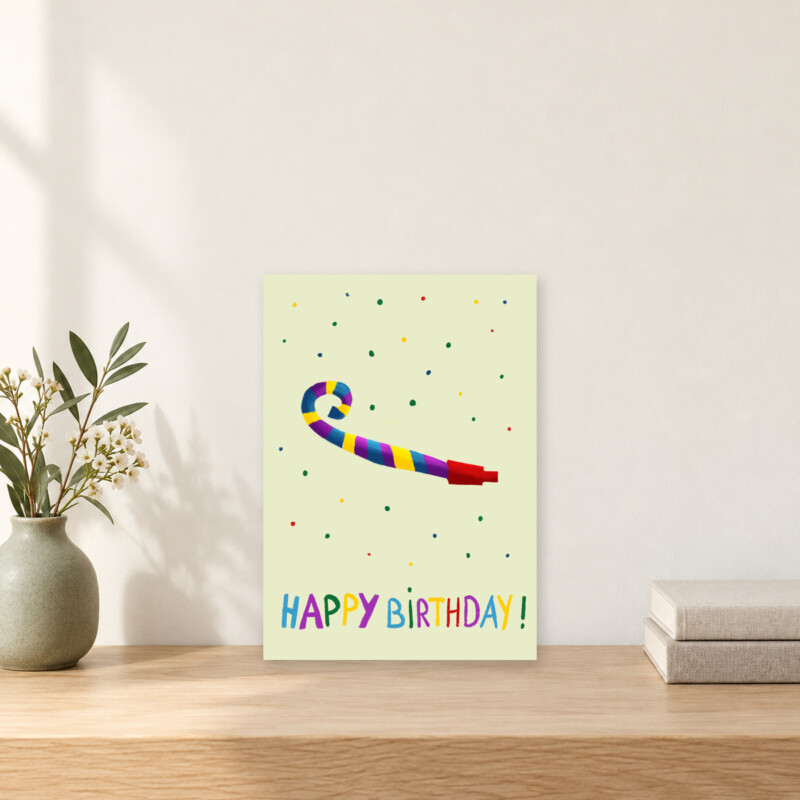 A6-verjaardagskaart met feesttoeter en confetti op een houten kastje, naast een vaasje en boeken in een licht interieur.